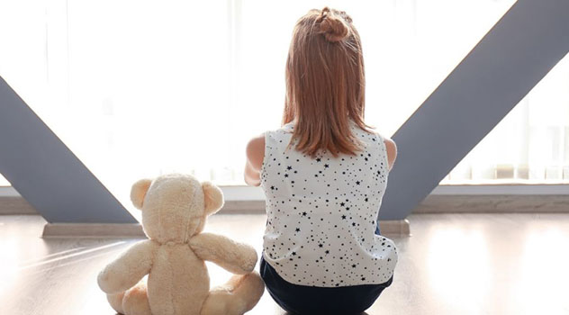 Una bambina seduta di spalle con le braccia incrociate seduta vicino ad un orsetto di peluche anch'esso girato di spalle