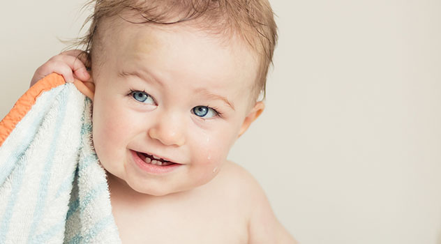 Un bambino piccolo con gli occhi azzurri dopo il bagnetto mentre si pulisce l'orecchio destro con un asciugamano