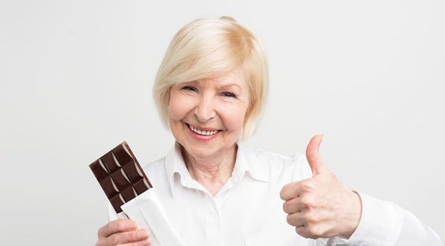 Donna anziana sorridente che mantiene una tavoletta di cioccolato con la mano destra ed ha il pollice sinistro alzato in segno di approvazione