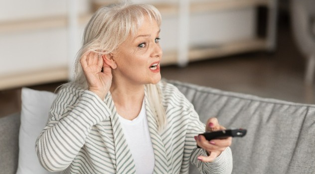 Donna seduta sul divano con un telecomando in mano e l'altra mano posizionata in prossimità dell'orecchio per sentire meglio