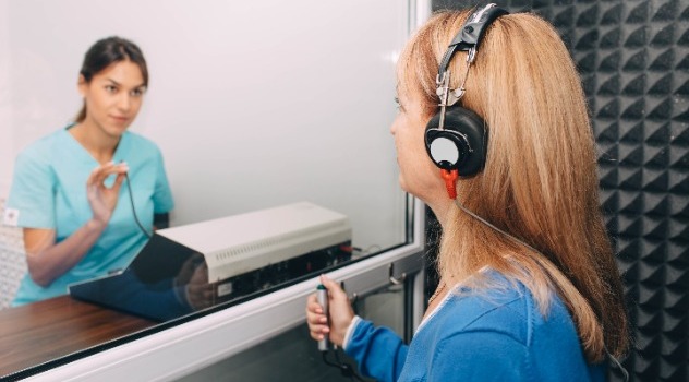 Una donna che sta eseguendo esami audiometrici indossando un paio di cuffie all'interno di una cabina insonorizzata mentre fuori la vetrata c'è una giovane dottoressa audiometrista in camice azzurro