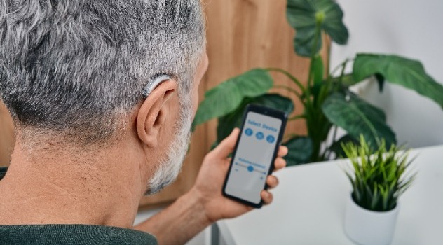 Immagine di spalle di un signore che indossa un apparecchio acustico retroauricolare dietro l'orecchio destro e mantiene con la mano sinistra uno smartphone