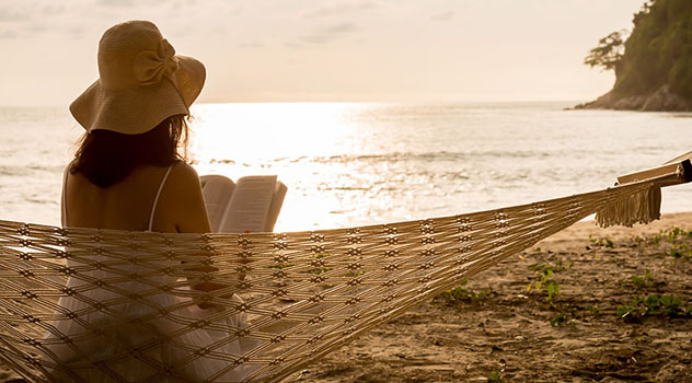 Ragazza in spiaggia seduta su un'amaca che legge un libro rivolta verso il mare durante il tramonto