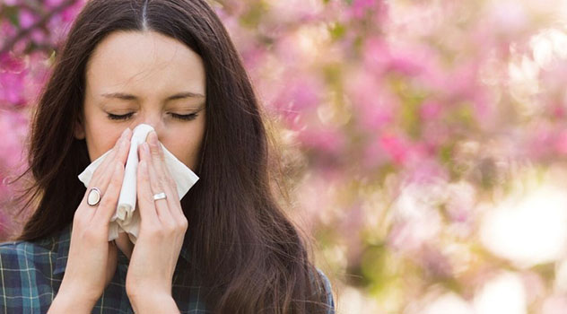 Ragazza con gli occhi chiusi mentre si soffia il naso, sullo sfondo un albero con fiori sfocato