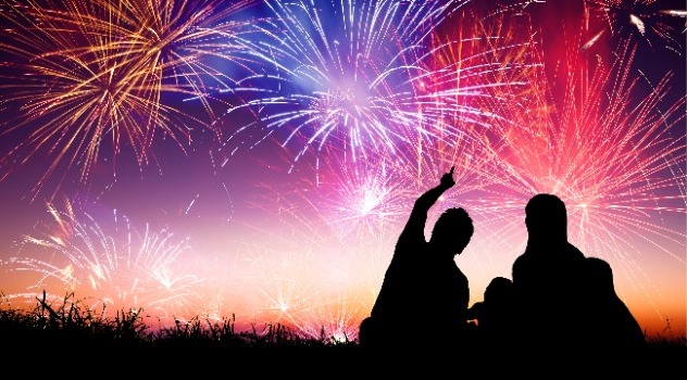 Silhouette scura di una famiglia distesa sul prato che ammira i fuochi d'artificio colorati su tonalità violacea mentre il padre indica lo spettacolo pirotecnico ai figli
