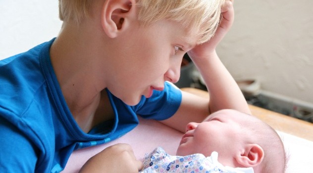 Un bambino con i capelli biondi in età scolare che parla ad un neonato mentre appoggiato con il gomito sul lettino dove sta-sdraiato il piccolo