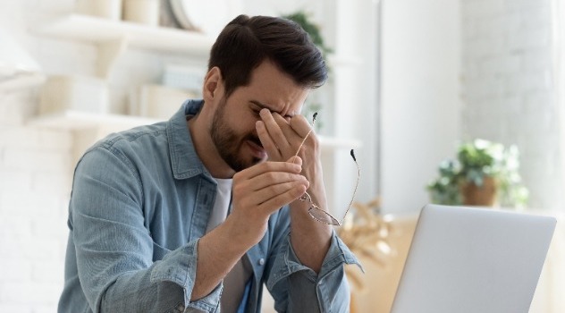 Uomo davanti al computer con espressione sofferente che mantiene gli occhiali con una mano e si preme sugli occhi con l'altra